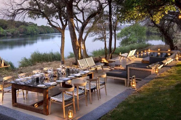 A long dining table laid beside the water under the trees, lit by lanterns at dusk